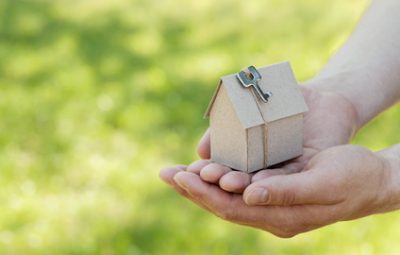 Male hands hold of cardboard house with key against natural green bokeh background. Building, loan, housewarming, insurance, real estate or buying a new home c